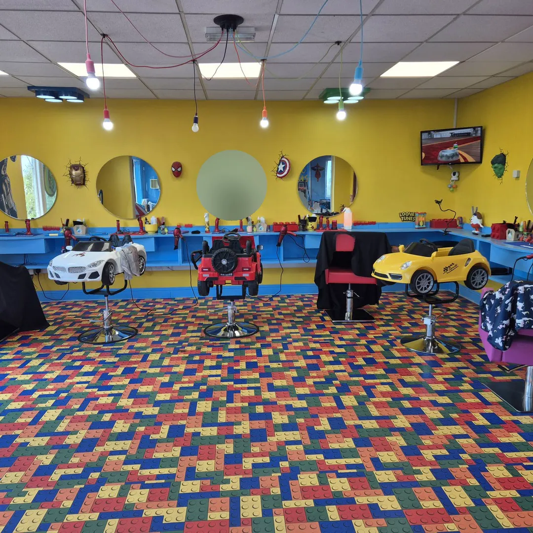 The barber floor: yellow walls, round mirrors, superhero masks and toy-car barber chairs on a colourful Lego-brick carpet.