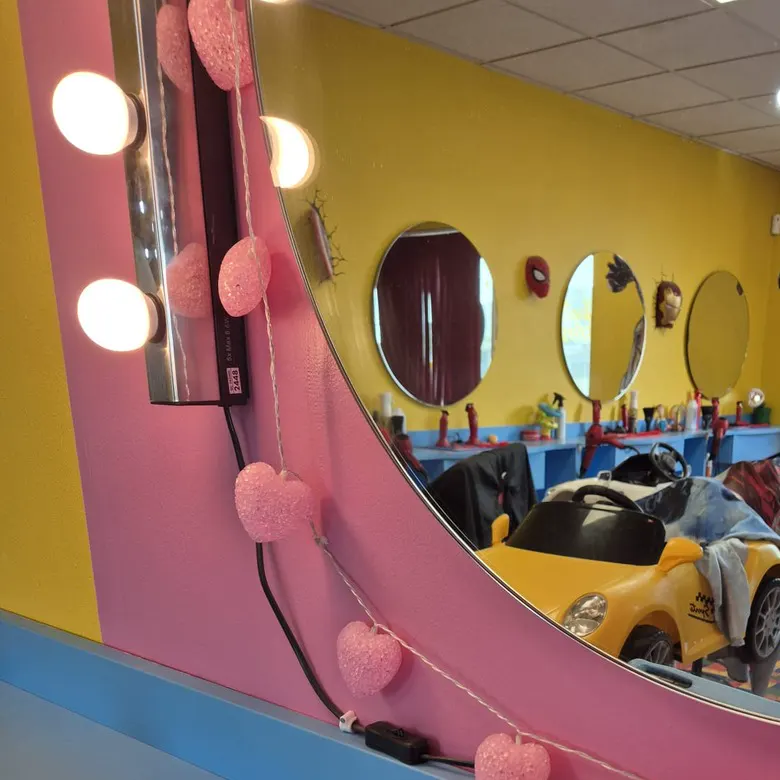 A pink mirror with light bulbs and heart lights, reflecting a yellow toy-car chair and round mirrors.